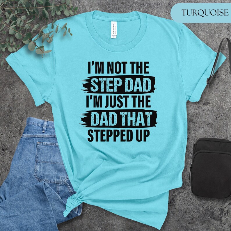 Puede incluir: Camiseta turquesa con el texto negro "I'M NOT THE STEP DAD I'M JUST THE DAD THAT STEPPED UP". La camiseta es de color azul claro y est&aacute; doblada en la parte inferior. Se ven unos vaqueros azules a la izquierda.