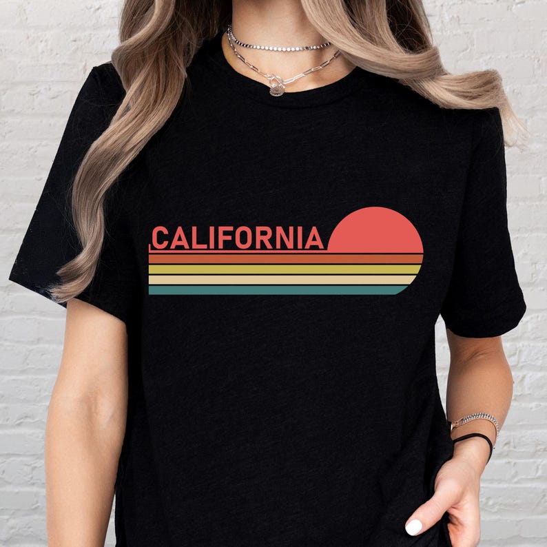 May include: Black t-shirt featuring the word "CALIFORNIA" in a retro font, above a horizontal design of orange, yellow, teal, and blue stripes, and a red circle. The design evokes a vintage sunset.