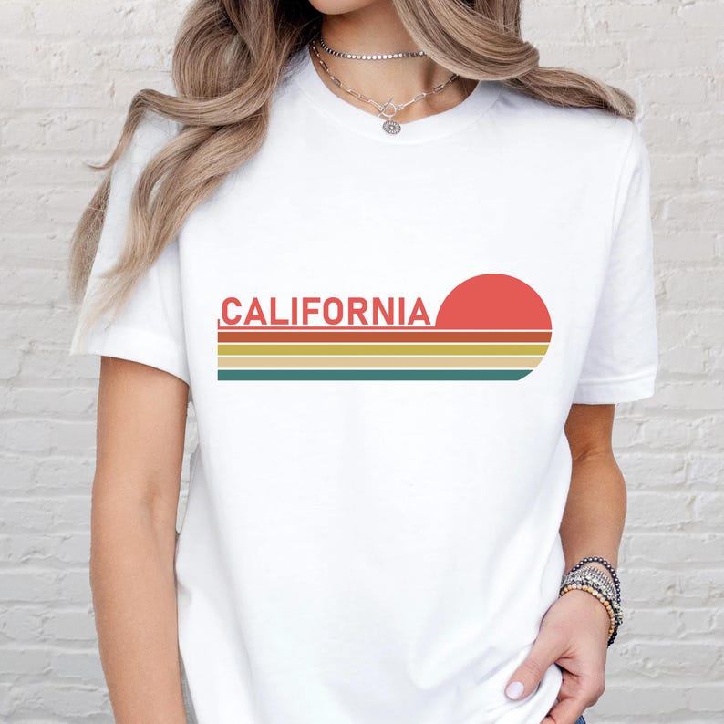 May include: White t-shirt with the word "CALIFORNIA" in red, above a retro sunset design with horizontal stripes in yellow, orange, and green. The shirt is a classic crew neck style.