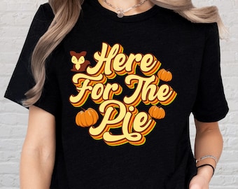 May include: Black t-shirt with the text "Here For The Pie" in a retro font, featuring pumpkins and a fox illustration. The design is in shades of yellow, orange, and brown.