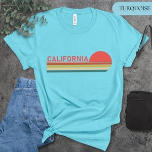May include: Turquoise t-shirt with the word "CALIFORNIA" in orange letters and a retro sunset design. The sunset features a red sun and horizontal stripes in orange, yellow, and green. The shirt is made of a soft material.