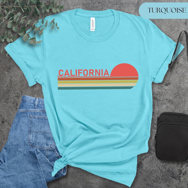 May include: Turquoise t-shirt with the word "CALIFORNIA" in orange letters and a retro sunset design. The sunset features a red sun and horizontal stripes in orange, yellow, and green. The shirt is made of a soft material.