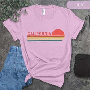 May include: Lilac-colored t-shirt with the word "CALIFORNIA" in red letters and a retro sunset design. The design includes a red sun and horizontal stripes in yellow, orange, and teal. The shirt is paired with blue jeans.