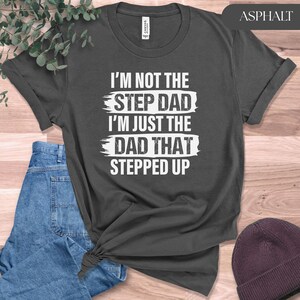 Puede incluir: Camiseta color asfalto con el texto blanco "I'M NOT THE STEP DAD I'M JUST THE DAD THAT STEPPED UP". La camiseta tiene cuello redondo y mangas enrolladas. La imagen incluye vaqueros azules y un gorro granate.