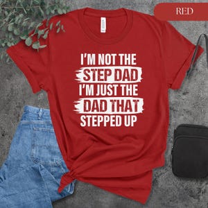 Puede incluir: Camiseta roja con texto blanco: "I'M NOT THE STEP DAD I'M JUST THE DAD THAT STEPPED UP." La camiseta se combina con vaqueros azules y una bolsa negra. La palabra "RED" est&aacute; en un rect&aacute;ngulo rojo en la esquina superior derecha.