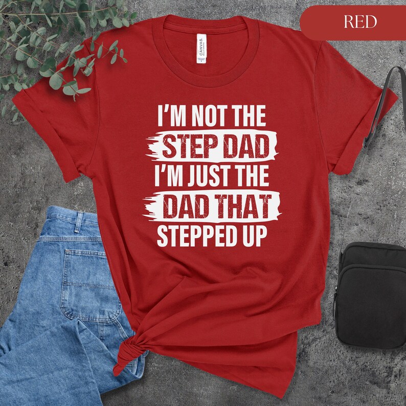 Puede incluir: Camiseta roja con texto blanco: "I'M NOT THE STEP DAD I'M JUST THE DAD THAT STEPPED UP." La camiseta se combina con vaqueros azules y una bolsa negra. La palabra "RED" est&aacute; en un rect&aacute;ngulo rojo en la esquina superior derecha.