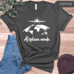 May include: Asphalt-colored t-shirt featuring a white graphic of an airplane above a world map, with the words "Airplane mode" in a handwritten font. The shirt is folded and tied at the bottom, with a pair of blue jeans and a maroon beanie in the background.