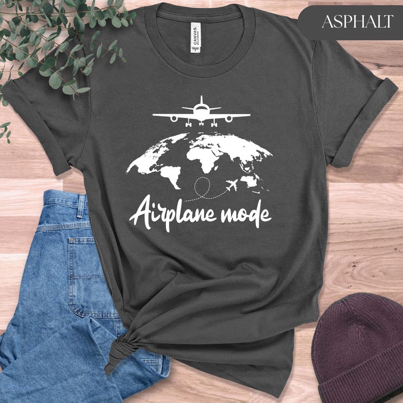 May include: Asphalt-colored t-shirt featuring a white graphic of an airplane above a world map, with the words "Airplane mode" in a handwritten font. The shirt is folded and tied at the bottom, with a pair of blue jeans and a maroon beanie in the background.