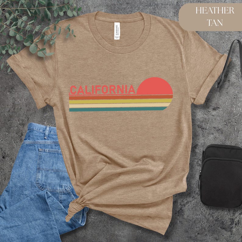 May include: Heather tan t-shirt with the word "CALIFORNIA" in coral letters and a retro sunset design. The design includes a red sun and horizontal stripes in yellow, teal, and orange. The shirt is made of soft material.