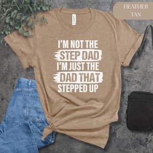 Puede incluir: Camiseta de color marr&oacute;n jaspeado con el texto blanco "I'M NOT THE STEP DAD I'M JUST THE DAD THAT STEPPED UP". La camiseta se combina con unos vaqueros azules y un bolso negro.