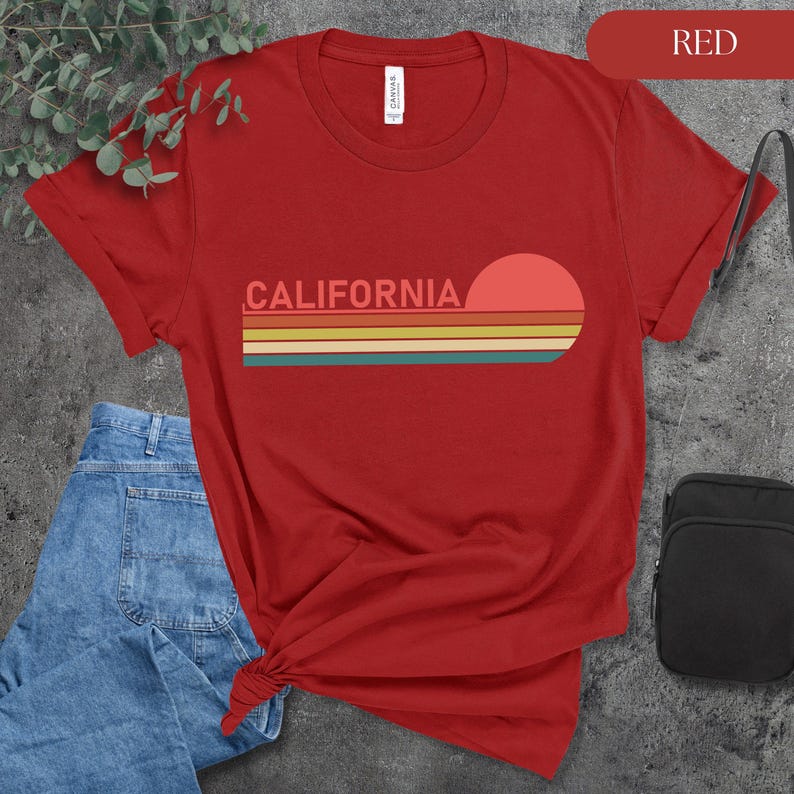 May include: Red t-shirt with the word "CALIFORNIA" in a retro font, above a horizontal design of colorful stripes and a setting sun. The shirt is paired with blue jeans and a black bag.