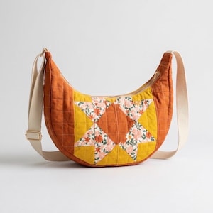 Puede incluir: Bolso bandolera acolchado en forma de media luna en tonos naranja, amarillo y estampado floral. El bolso presenta un diseño de estrella en la parte delantera, una correa ajustable beige y una cremallera dorada.