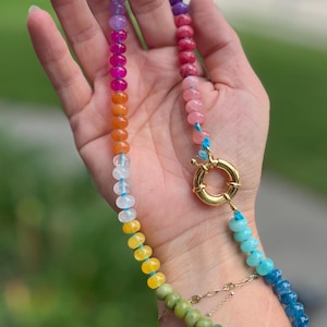 May include: A colorful beaded necklace with a rainbow of colors, including red, orange, yellow, green, blue, and purple. The necklace has a gold clasp and is held in a hand.