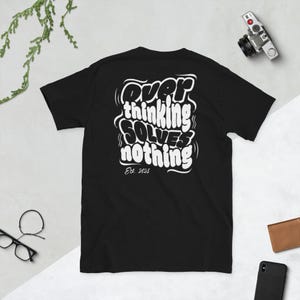 Puede incluir: Camiseta negra con el texto blanco "Overthinking Solves Nothing" en una fuente estilizada. El texto "Est. 2025" está debajo. La camiseta se muestra sobre un fondo claro.