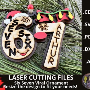 May include: Wooden Christmas ornament featuring the numbers "6" and "7" with festive designs. The number "6" has a Santa hat and the word "SIX" inside. The number "7" has the name "ARTHUR" on it. The ornament is on a bed of pine needles.