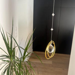 May include: A hanging crystal suncatcher with a clear, faceted crystal pendant suspended within two gold-colored rings. The suncatcher hangs from a thin, silver-colored string with three smaller crystal beads.