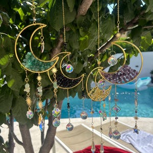 Può includere: Acchiappasole a forma di mezzaluna in metallo color oro con vari accenti di cristalli e pietre preziose. Ogni mezzaluna è riempita con pietre di diversi colori e pende da una catena con cristalli pendenti e ciondoli a forma di stella. Gli acchiappasole sono appesi a un albero.