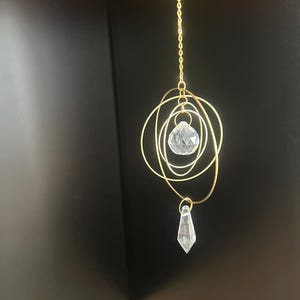 Peut inclure: Ornement décoratif suspendu, composé d'une chaîne et de cercles en fil de couleur or. Un cristal facetté est suspendu en bas, avec une sphère de cristal plus grande suspendue au centre des cercles. L'ornement est conçu pour capter la lumière.