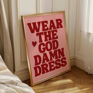 Könnte beinhalten: Ein gerahmter rosa Kunstdruck mit dem Text "WEAR THE GOD DAMN DRESS" in fetten roten Buchstaben. Ein rotes Herz befindet sich zwischen "WEAR" und "THE". Der Rahmen ist goldfarben.