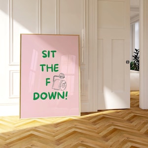 May include: A framed pink poster with the text "SIT THE F DOWN!" in green, featuring a toilet paper roll illustration. The poster is in a gold frame and leans against a wall with a wooden floor.