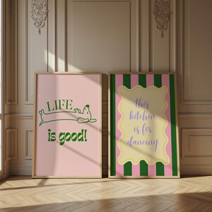 Peut inclure: Deux affiches encadr&eacute;es. L'affiche de gauche a un fond rose avec du texte vert et une illustration de teckel qui dit "LIFE is good!". L'affiche de droite a un fond ray&eacute; rose et vert avec une forme jaune et le texte "this kitchen is for dancing".