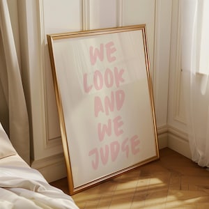Könnte beinhalten: Ein gerahmter Kunstdruck mit den Worten "WE LOOK AND WE JUDGE" in rosa Schrift. Der Rahmen ist goldfarben und der Hintergrund ist hellcremefarben. Das Kunstwerk lehnt an einer Wand.