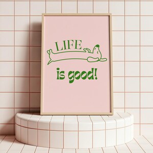 Peut inclure: Une affiche encadr&eacute;e avec un fond rose clair et les mots "LIFE is good!" en vert. Un teckel vert de dessin anim&eacute; se pr&eacute;lassant au-dessus des mots. Le cadre est de couleur bois clair. L'affiche est expos&eacute;e sur un pi&eacute;destal blanc et arrondi.