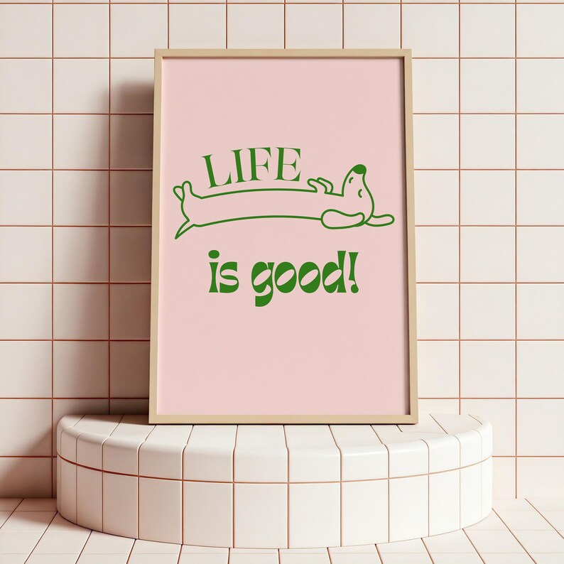 Peut inclure: Une affiche encadr&eacute;e avec un fond rose clair et les mots "LIFE is good!" en vert. Un teckel vert de dessin anim&eacute; se pr&eacute;lassant au-dessus des mots. Le cadre est de couleur bois clair. L'affiche est expos&eacute;e sur un pi&eacute;destal blanc et arrondi.