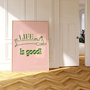 Peut inclure: Affiche rose encadr&eacute;e avec les mots "LIFE is good!" en vert, avec une illustration de teckel. L'affiche est expos&eacute;e dans une pi&egrave;ce avec un sol en bois et des murs blancs.