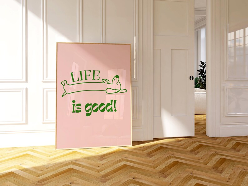 Peut inclure: Affiche rose encadr&eacute;e avec les mots "LIFE is good!" en vert, avec une illustration de teckel. L'affiche est expos&eacute;e dans une pi&egrave;ce avec un sol en bois et des murs blancs.