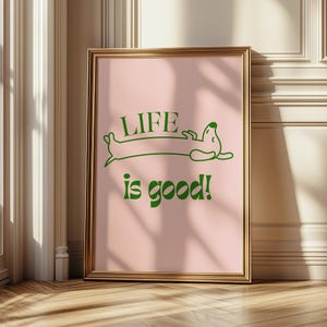 Peut inclure: Une affiche encadr&eacute;e avec un cadre dor&eacute;. L'impression pr&eacute;sente un fond rose avec une illustration de teckel vert et les mots "LIFE is good!" en vert. L'&oelig;uvre est expos&eacute;e sur un sol en bois.