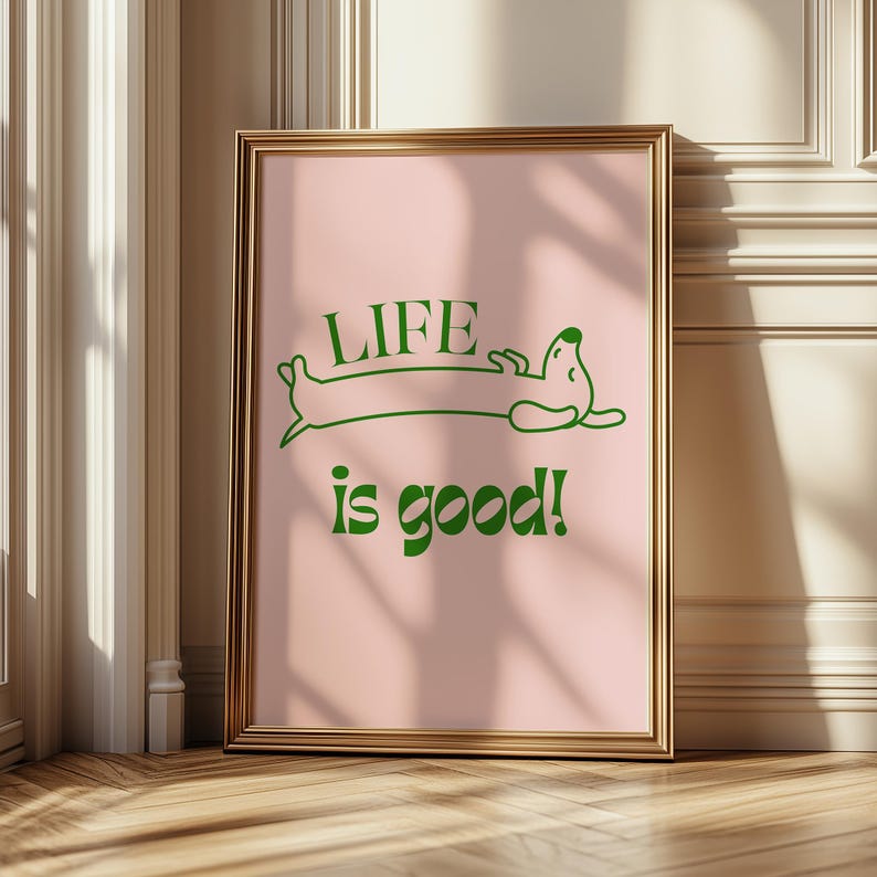 Peut inclure: Une affiche encadr&eacute;e avec un cadre dor&eacute;. L'impression pr&eacute;sente un fond rose avec une illustration de teckel vert et les mots "LIFE is good!" en vert. L'&oelig;uvre est expos&eacute;e sur un sol en bois.