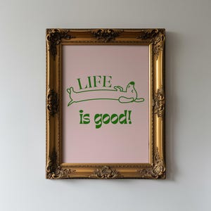 Peut inclure: Une affiche encadr&eacute;e dans un cadre dor&eacute; orn&eacute;. L'affiche a un fond rose clair avec du texte vert et une illustration de teckel. Le texte dit "LIFE is good!"