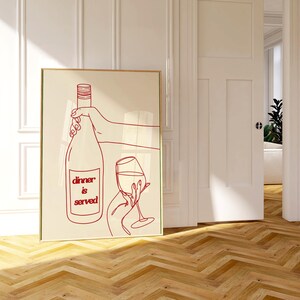 May include: A framed art print featuring a red line drawing of a wine bottle and glass, with the text "dinner is served." The artwork is set against a cream background, with a gold frame. The print is displayed on a wooden floor.