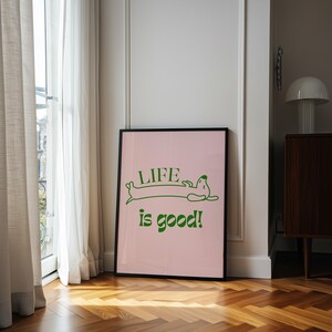 Peut inclure: Affiche encadr&eacute;e avec un fond rose et les mots "LIFE is good!" en vert. Le mot "LIFE" est au-dessus d'une illustration de teckel vert. L'affiche est dans un cadre noir et est appuy&eacute;e contre un mur blanc.