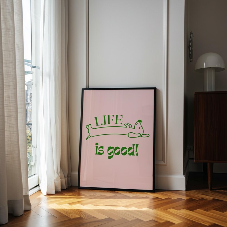 Peut inclure: Affiche encadr&eacute;e avec un fond rose et les mots "LIFE is good!" en vert. Le mot "LIFE" est au-dessus d'une illustration de teckel vert. L'affiche est dans un cadre noir et est appuy&eacute;e contre un mur blanc.