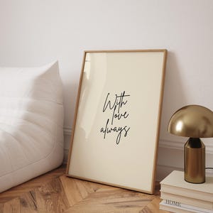 Könnte beinhalten: Ein gerahmter Druck mit den Worten "With love always" in eleganter Schrift. Das Kunstwerk hat einen cremefarbenen Hintergrund und einen hellbraunen Holzrahmen. Eine goldfarbene Lampe und Bücher befinden sich rechts, und ein weißer Sitzsack links.