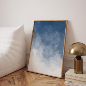 Könnte beinhalten: Ein gerahmter Kunstdruck mit einem Aquarelldesign in Blau- und Weißtönen. Das Kunstwerk wird auf einem Holzboden präsentiert, der an einer weißen Wand lehnt. Eine goldene Lampe und Bücher sind ebenfalls zu sehen.