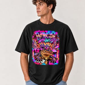 Puede incluir: Una camiseta negra de manga corta con un estampado gráfico vibrante de un caimán con gafas de sol y una cadena de oro. El diseño incluye un coche deportivo morado, palmeras y el texto "VICE WYNWOOD" en una fuente de colores neón.