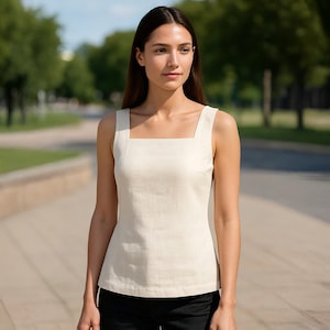 Op de afbeelding: Een crèmekleurige tanktop met een vierkante halslijn. De mouwloze top is gemaakt van een gladde stof en wordt gecombineerd met een zwarte broek. De achtergrond toont een vage buitenscène met bomen en een pad.