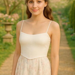 May include: A woman wearing a cream-colored dress with a floral print skirt and a square neckline. The dress has thin straps and a fitted bodice. The background is a garden with a path and trees.