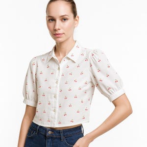 May include: White button-down shirt with a cherry print. The shirt has short, puffed sleeves and a collared neckline. The cherries are red with green stems, and the shirt is paired with blue jeans.