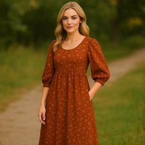 May include: A rust-colored dress with a floral pattern. The dress features a scoop neckline, three-quarter sleeves, and a gathered waist. It has pockets and falls to mid-calf length. The background is a blurred outdoor scene with green foliage and a path.
