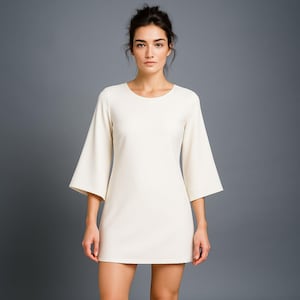 Op de afbeelding: Crèmekleurige, korte jurk met een ronde halslijn en uitlopende driekwartmouwen. De jurk is knielang en heeft een eenvoudig, elegant ontwerp. Het model staat voor een grijze achtergrond.