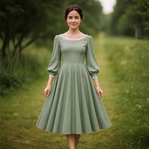 Pode incluir: Um vestido verde sálvia com decote redondo e mangas compridas bufantes. O vestido apresenta um corpete justo e uma saia rodada, com delicados acabamentos em renda no decote e nos punhos. A mulher está em pé num campo gramado.