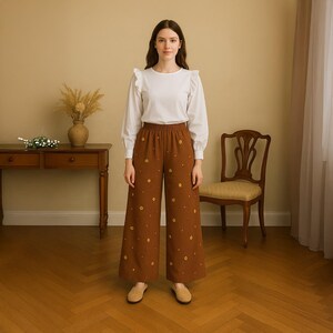 May include: A person wearing a white blouse with frill details and brown wide-leg trousers. The trousers feature a yellow floral pattern. The outfit is completed with tan flats. The background includes a wooden table, chair, and a vase with dried plants.