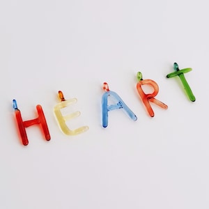 Puede incluir: Letras de vidrio de colores que deletrean la palabra "HEART". Las letras son rojas, amarillas, azules y verdes. Cada letra tiene un peque&ntilde;o bucle en la parte superior, lo que sugiere que podr&iacute;an usarse como amuletos o colgantes.
