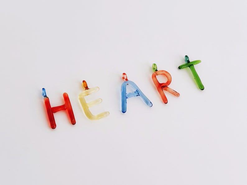 Puede incluir: Letras de vidrio de colores que deletrean la palabra "HEART". Las letras son rojas, amarillas, azules y verdes. Cada letra tiene un peque&ntilde;o bucle en la parte superior, lo que sugiere que podr&iacute;an usarse como amuletos o colgantes.