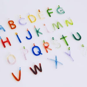 Puede incluir: Una colecci&oacute;n de colgantes de letras del alfabeto de vidrio de colores. Cada letra es de un color diferente, incluyendo azul, naranja, amarillo, verde y morado. Las letras tienen peque&ntilde;os bucles met&aacute;licos para sujetarlas a joyas u otras manualidades.