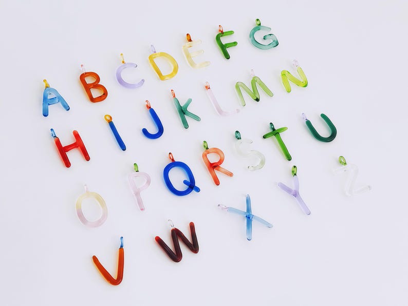 Puede incluir: Una colecci&oacute;n de colgantes de letras del alfabeto de vidrio de colores. Cada letra es de un color diferente, incluyendo azul, naranja, amarillo, verde y morado. Las letras tienen peque&ntilde;os bucles met&aacute;licos para sujetarlas a joyas u otras manualidades.
